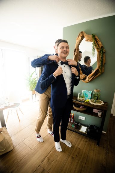 Bräutigam beim Getting Ready in Heilbronn – Trauzeuge hilft dem Bräutigam beim Anziehen des Hochzeitsanzugs, Nahaufnahme eleganter Details, 