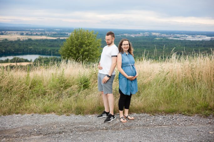 Schwangere Frau und Mann stehen nebeneinander auf einer Wiese mit Ausblick ins Grüne.