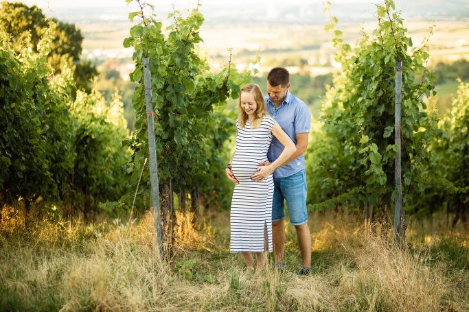 Schwangere Frau in gestreiftem Kleid steht in einem Weinberg, umarmt von einem Mann.