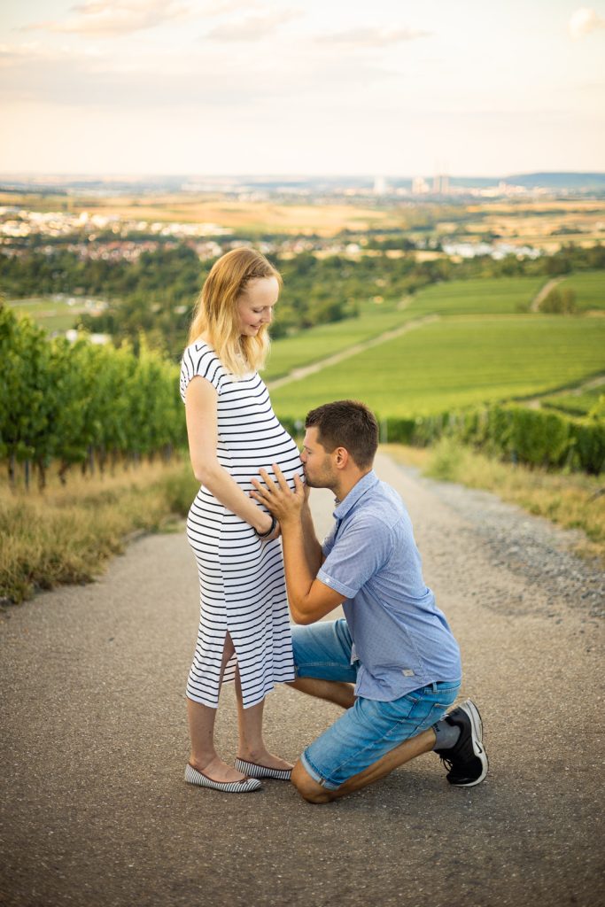 Ein Paar steht auf einem Weg; der Mann kniet und berührt den Bauch der schwangeren Frau.