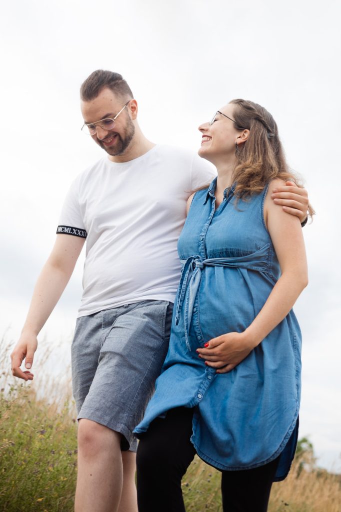 Paar in freier Natur, der Mann lächelt, die Frau trägt einen Bauch und hält ihn.