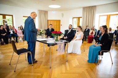 Eine Hochzeitszeremonie mit Gästen in festlicher Kleidung und einem Redner am Tisch.